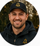 Portrait of a smiling light-skinned adult man wearing a dark baseball cap and dark work jacket.