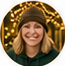 Circular portrait of a smiling woman in a knit hat, against warm decorative lighting.